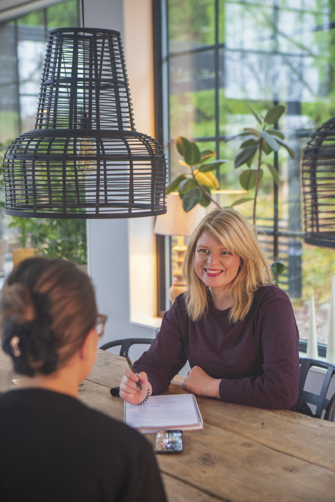 två kvinnor som sitter och samtalar vid ett bord. Den ena kvinnan visar något på ett papper.